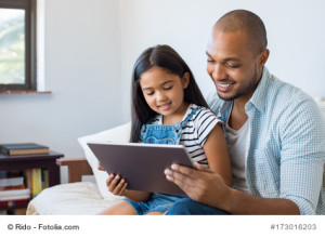 African father and smiling daughter sitting on sofa using digital tablet. Happy dad watching cartoon with his cute little girl. Cheerful father and daughter sitting on couch at home and playing with computer.